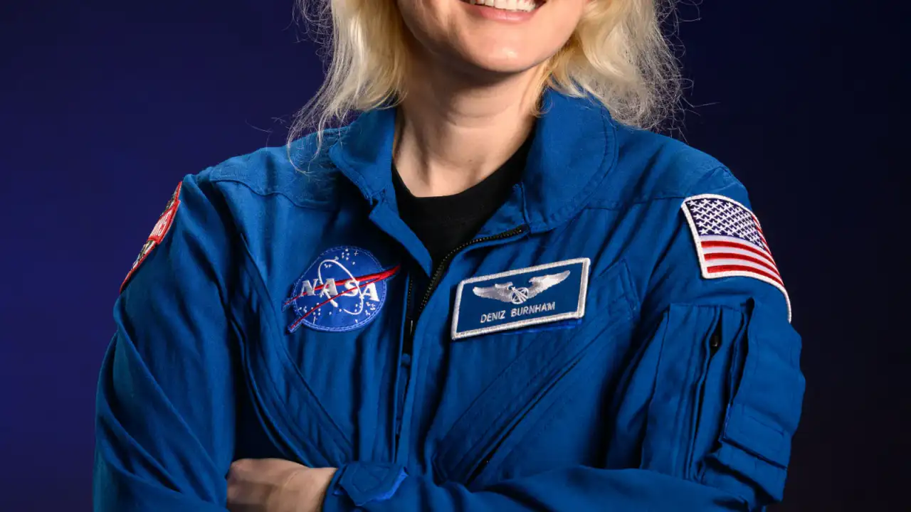 Astronauta Deniz Burnham con traje espacial frente a fondo de estación espacial