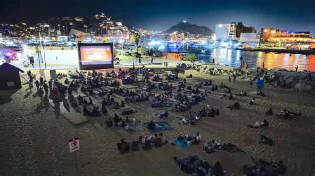 Familias sentadas en la arena de una playa viendo una película en una pantalla al aire libre durante la noche