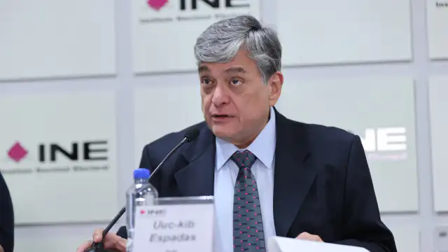 Boletas electorales sobre una mesa con el logo del INE al fondo