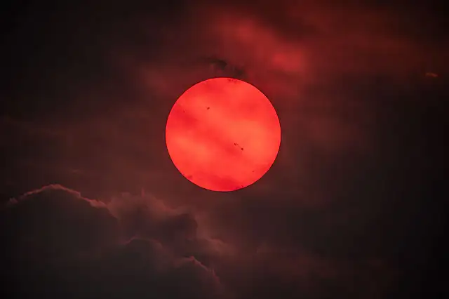 Fotografía del Sol de color rojo intenso debido al humo de incendios, con manchas solares visibles en la superficie.